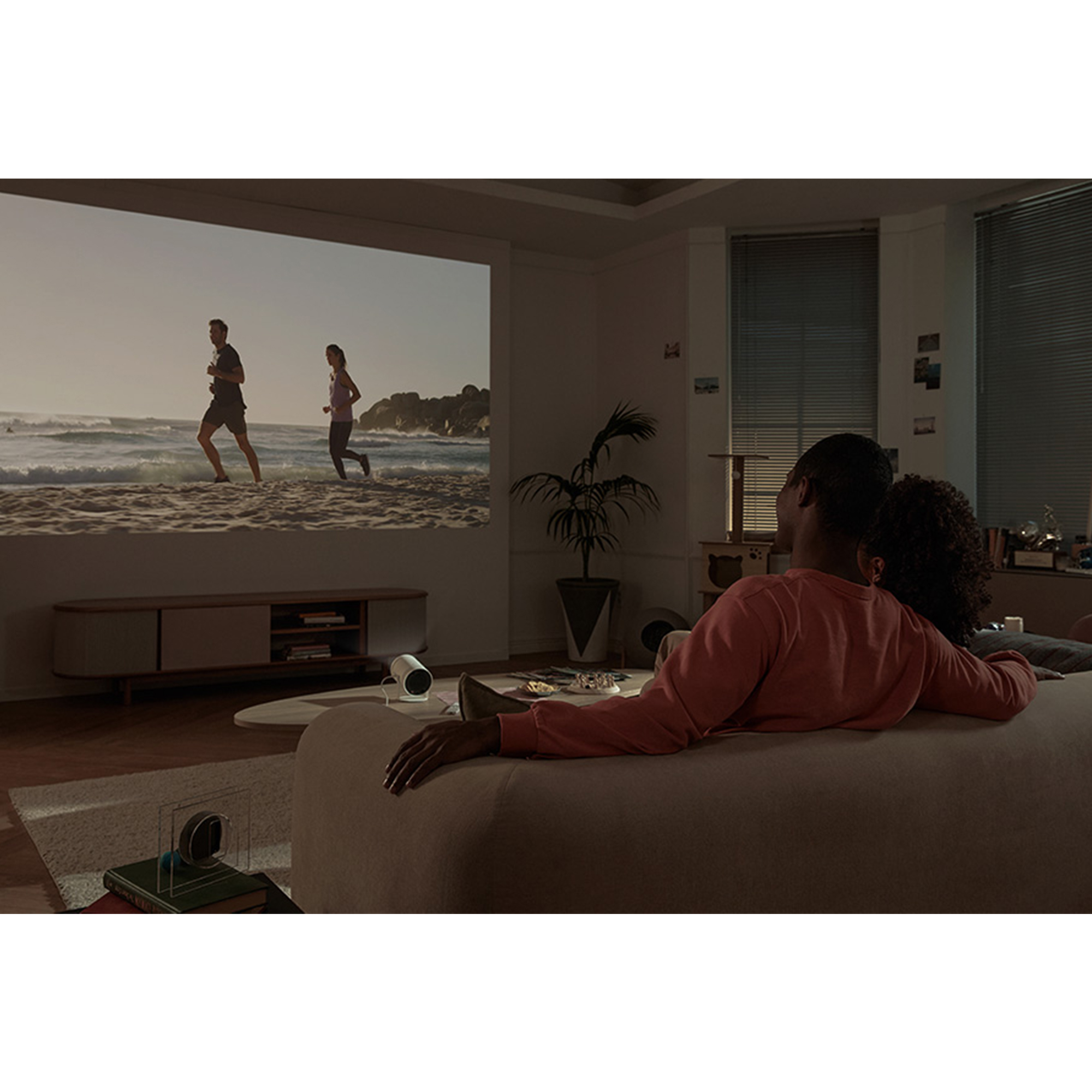 Dos personas ven una película proyectada en una pared. Se sientan en un sofá con un proyector y una mesa auxiliar.