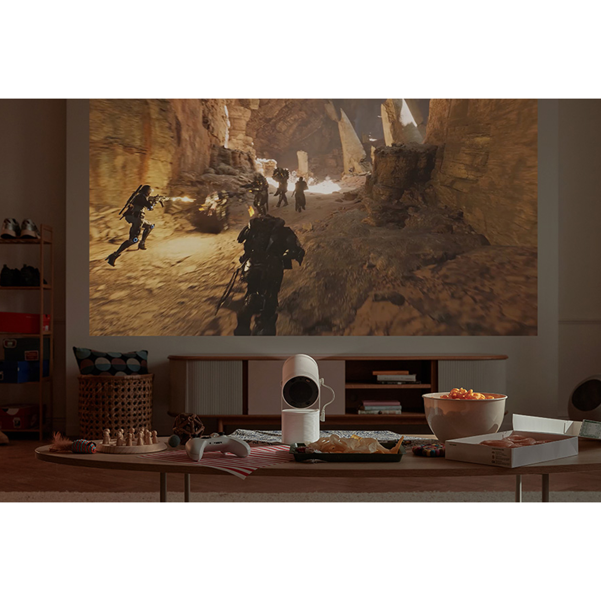 Un proyector blanco muestra una escena de videojuego en una pared, con aperitivos y una consola.