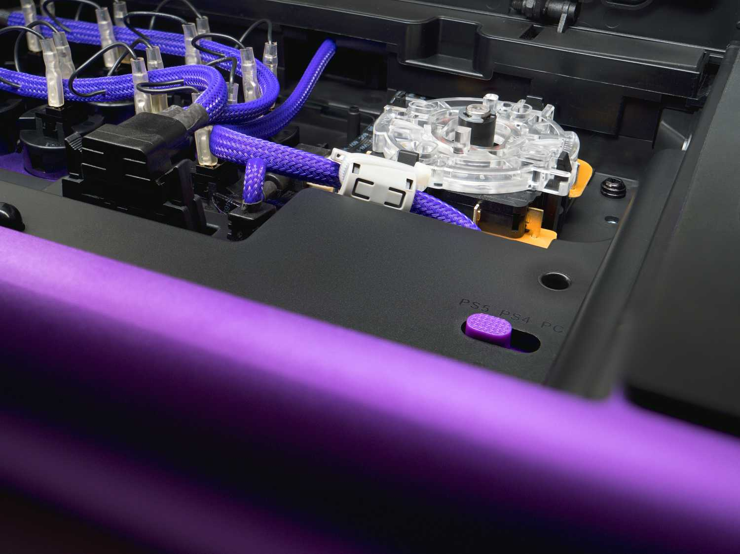 Primer plano del interior de una consola de juegos. Cables trenzados morados, un joystick transparente y un botón morado etiquetado PS5, PS4, PC son visibles.