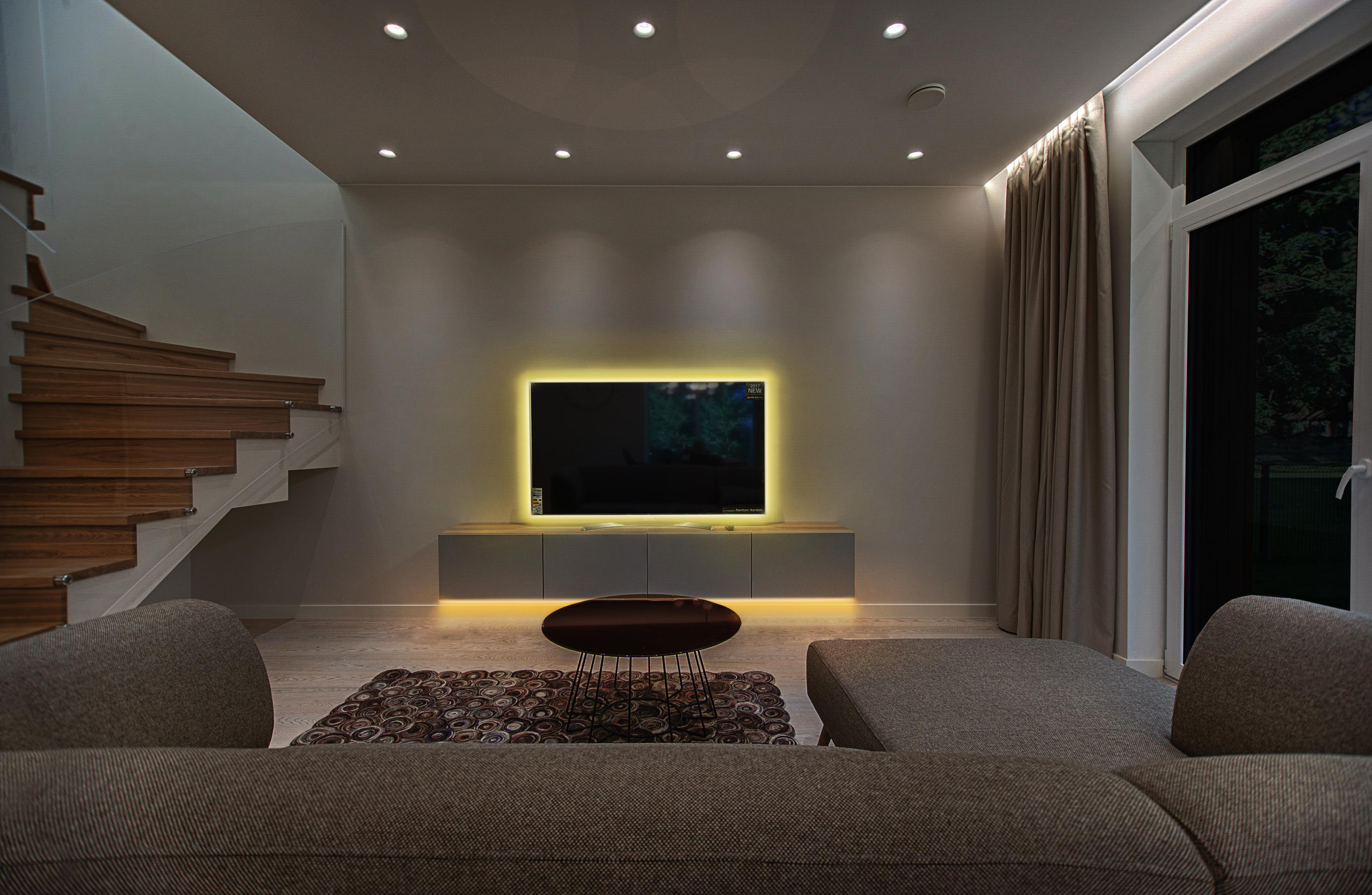 Modernes Wohnzimmer mit Fernseher an der Wand. Treppe und Sofa im Hintergrund. Warme Beleuchtung.