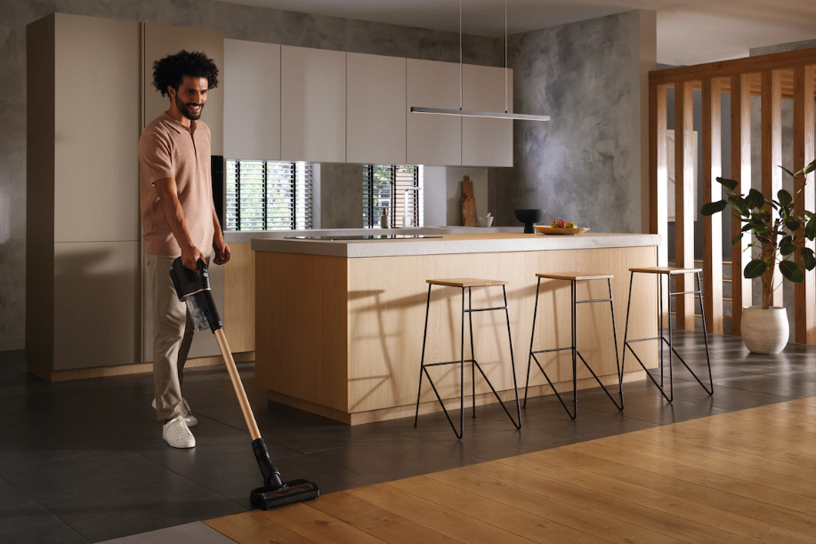 Mann, der einen Raum mit Holzboden saugt. Dahinter eine Küche mit Hockern und Schränken.