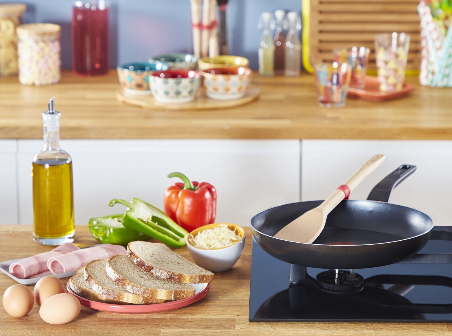 Scène de cuisine avec préparation des aliments. Poêle sur la cuisinière, légumes, œufs, pain et huile visibles.