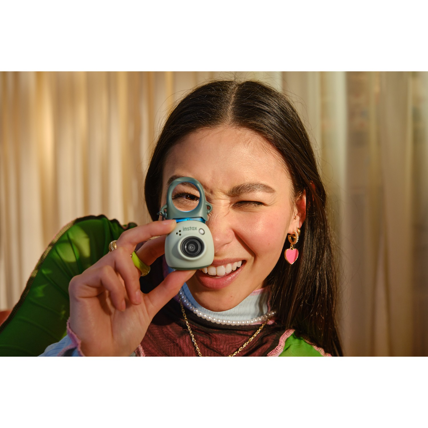 Vrouw knipoogt terwijl ze een kleine groene en blauwe camera vasthoudt. Ze draagt oorbellen en een parelketting.
