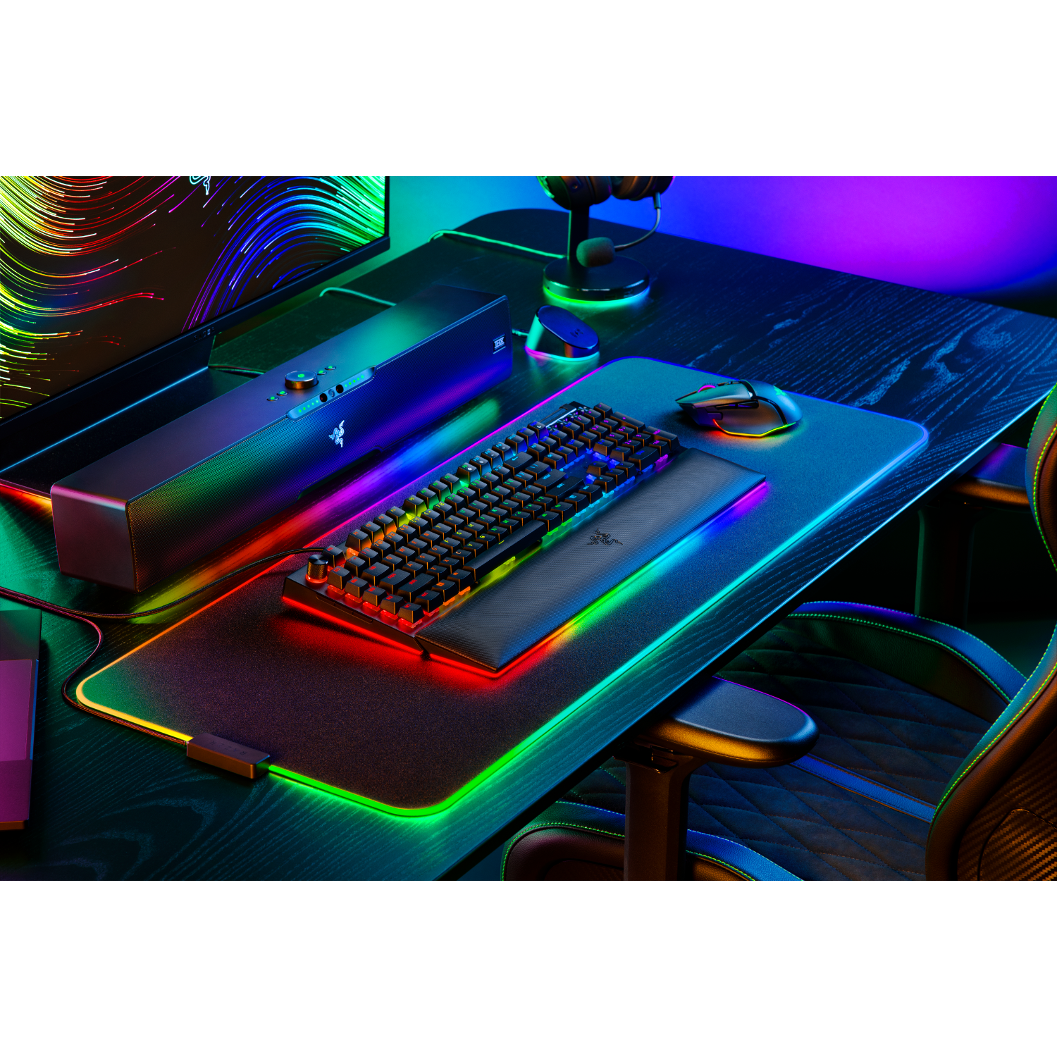 Gaming setup met een zwart toetsenbord en muis, kleurrijke lichten en een soundbar op een zwart bureau.
