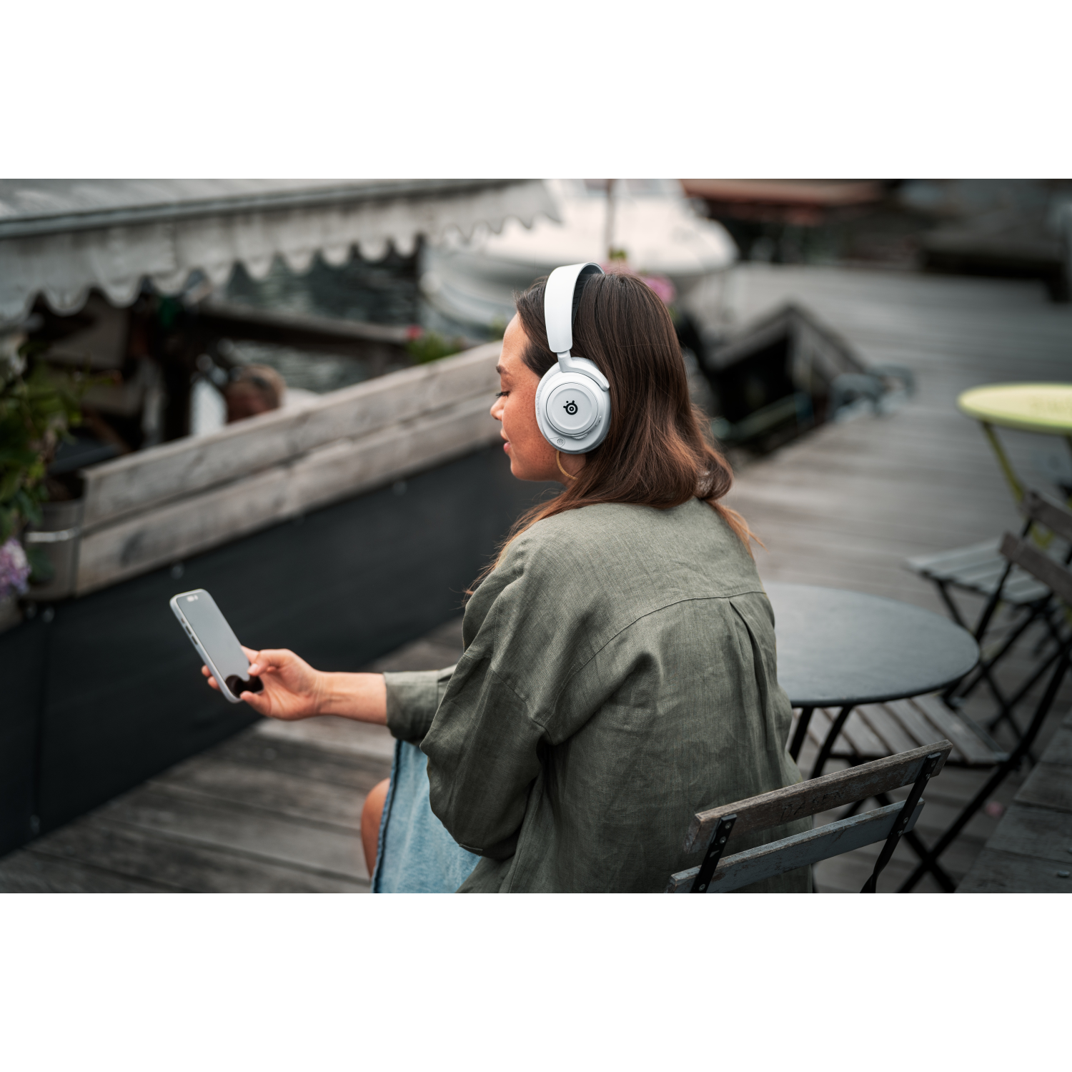 A woman with white headphones, holding a phone, sitting on a wooden dock. Dark background.