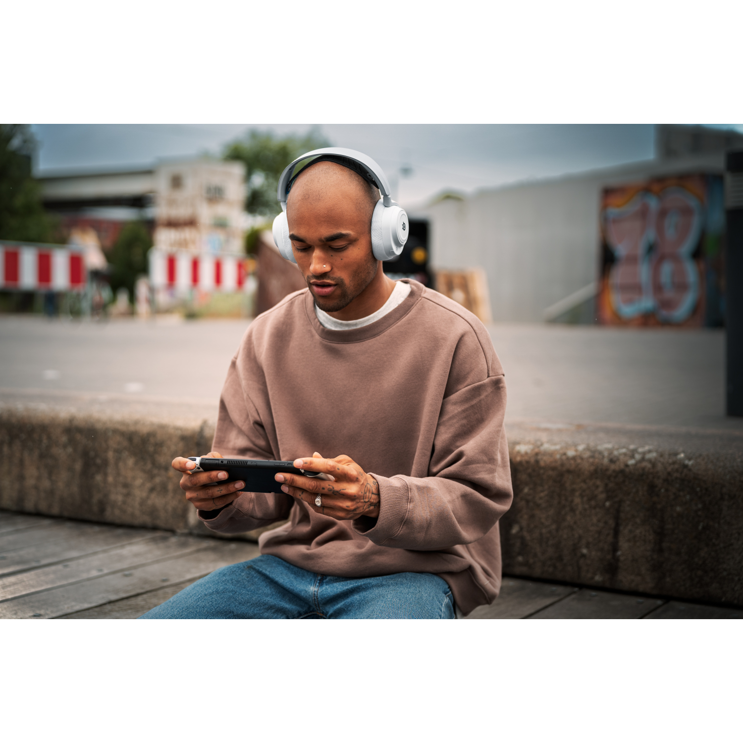 A man wearing headphones plays a game on a handheld device. Sitting on a wooden surface outside.