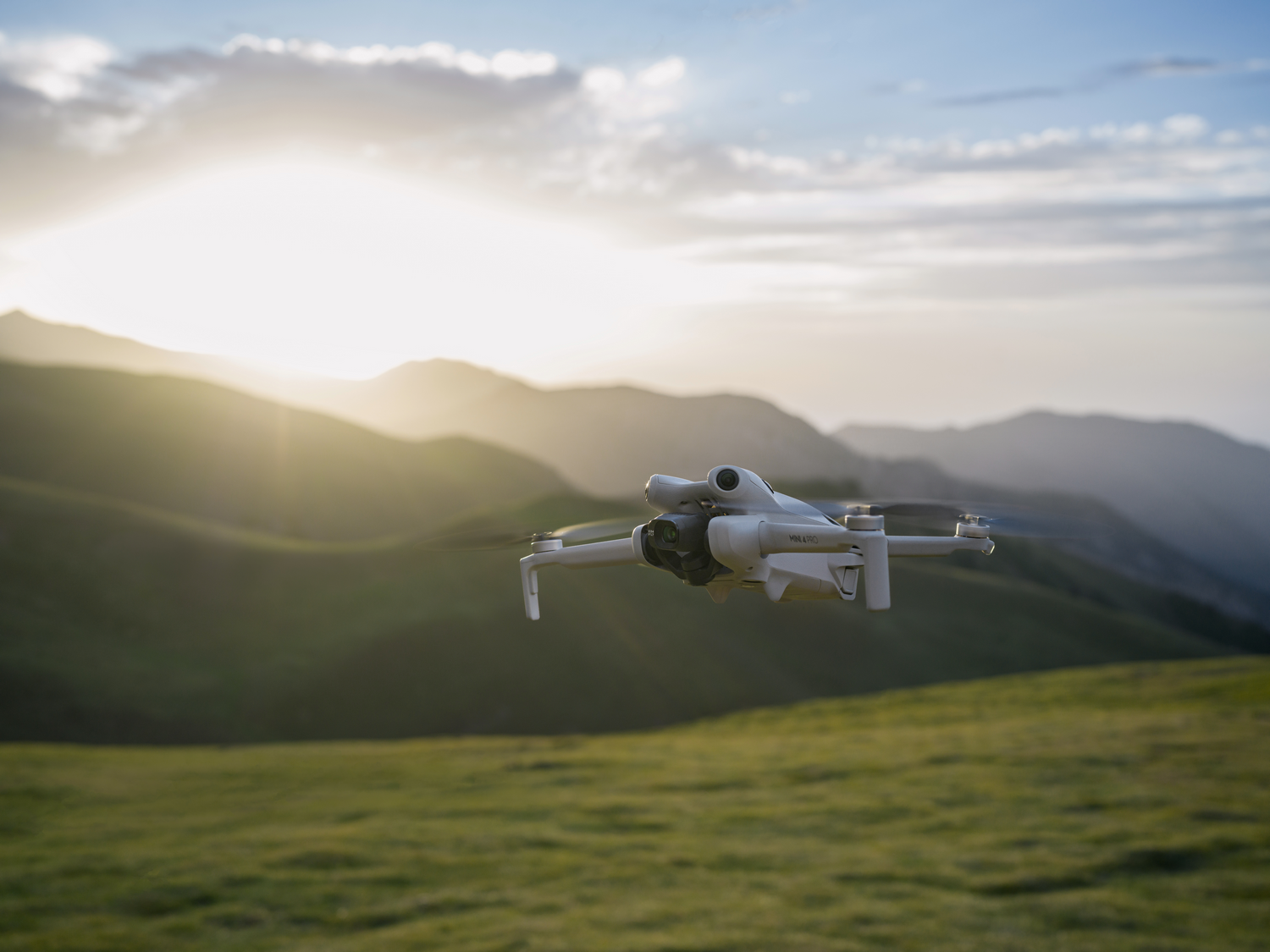 Un dron blanco volando sobre colinas verdes con el sol brillando intensamente al fondo.