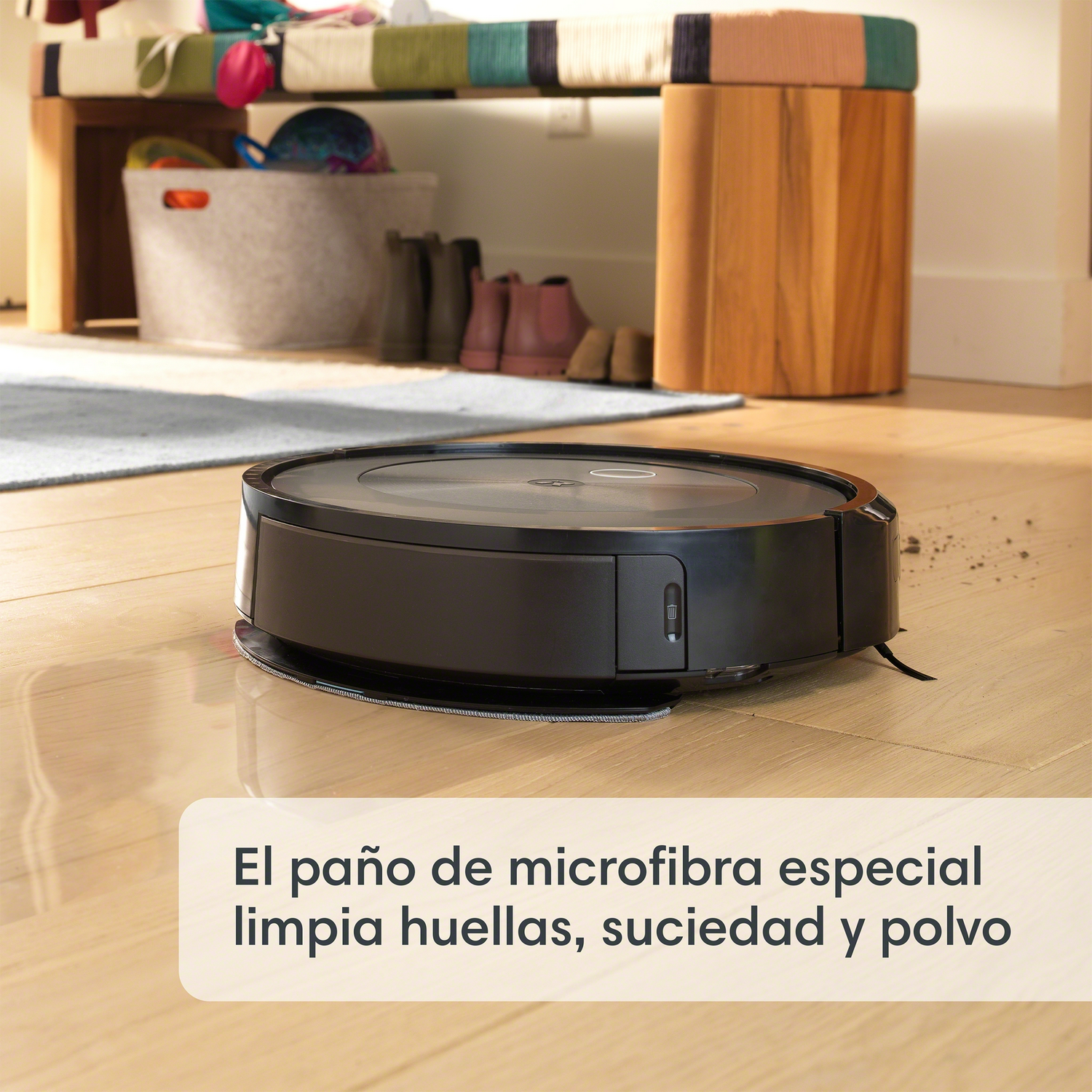 Aspiradora robot negra en el suelo. El fondo muestra un banco, zapatos y una puerta.