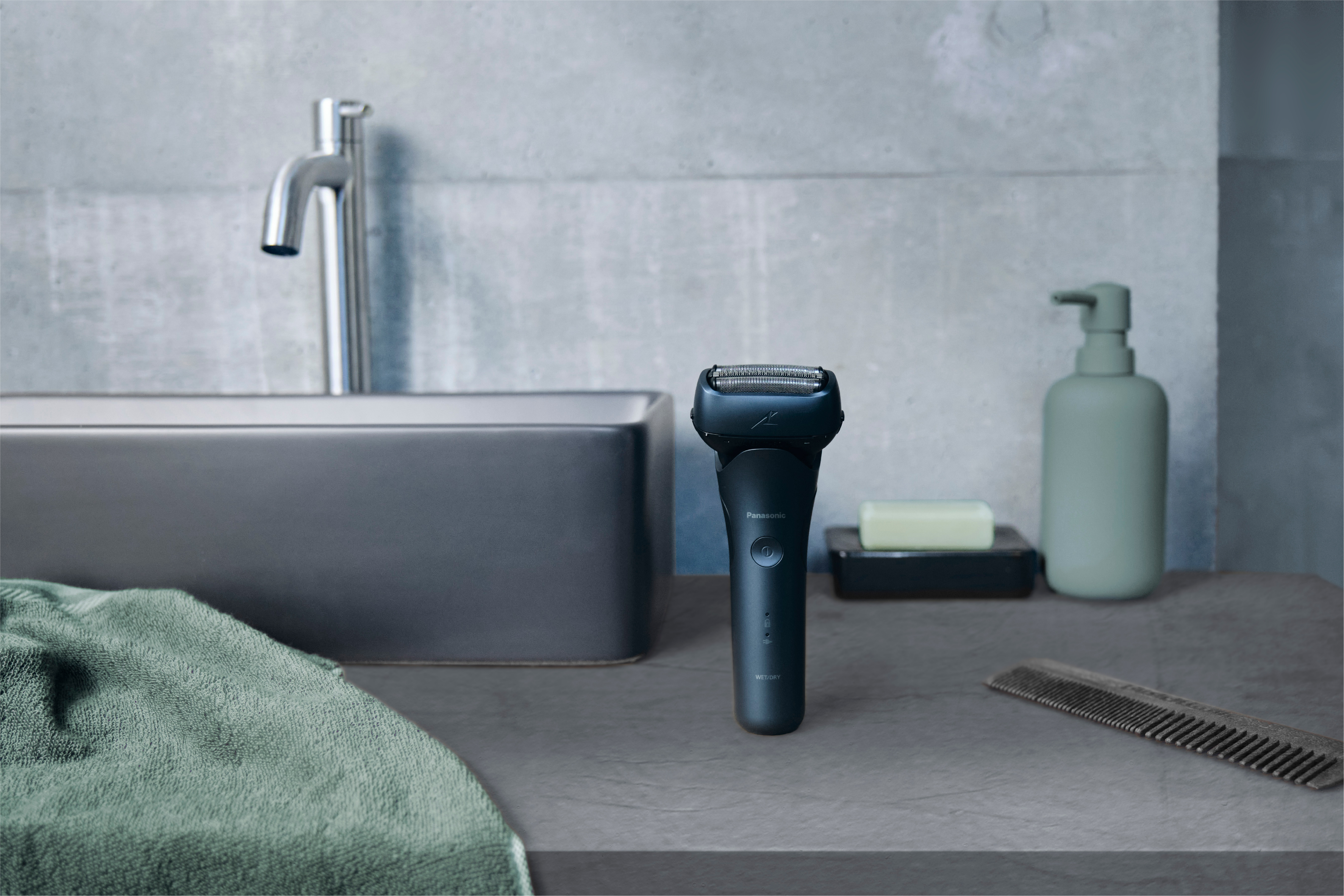 Elektrischer Rasierer auf Arbeitsplatte mit Waschbecken, Handtuch, Seife und Kamm.