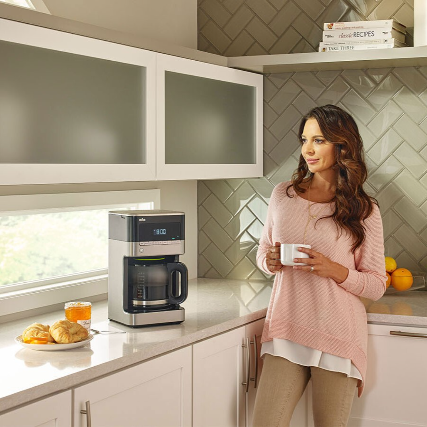 Woman with coffee by coffee maker, kitchen setting. Croissants and oranges on table.