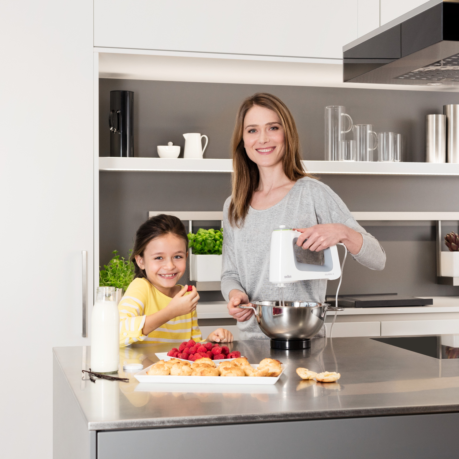 A woman and a child are in a kitchen. The woman is using a mixer. Raspberries and pastries are on a plate.