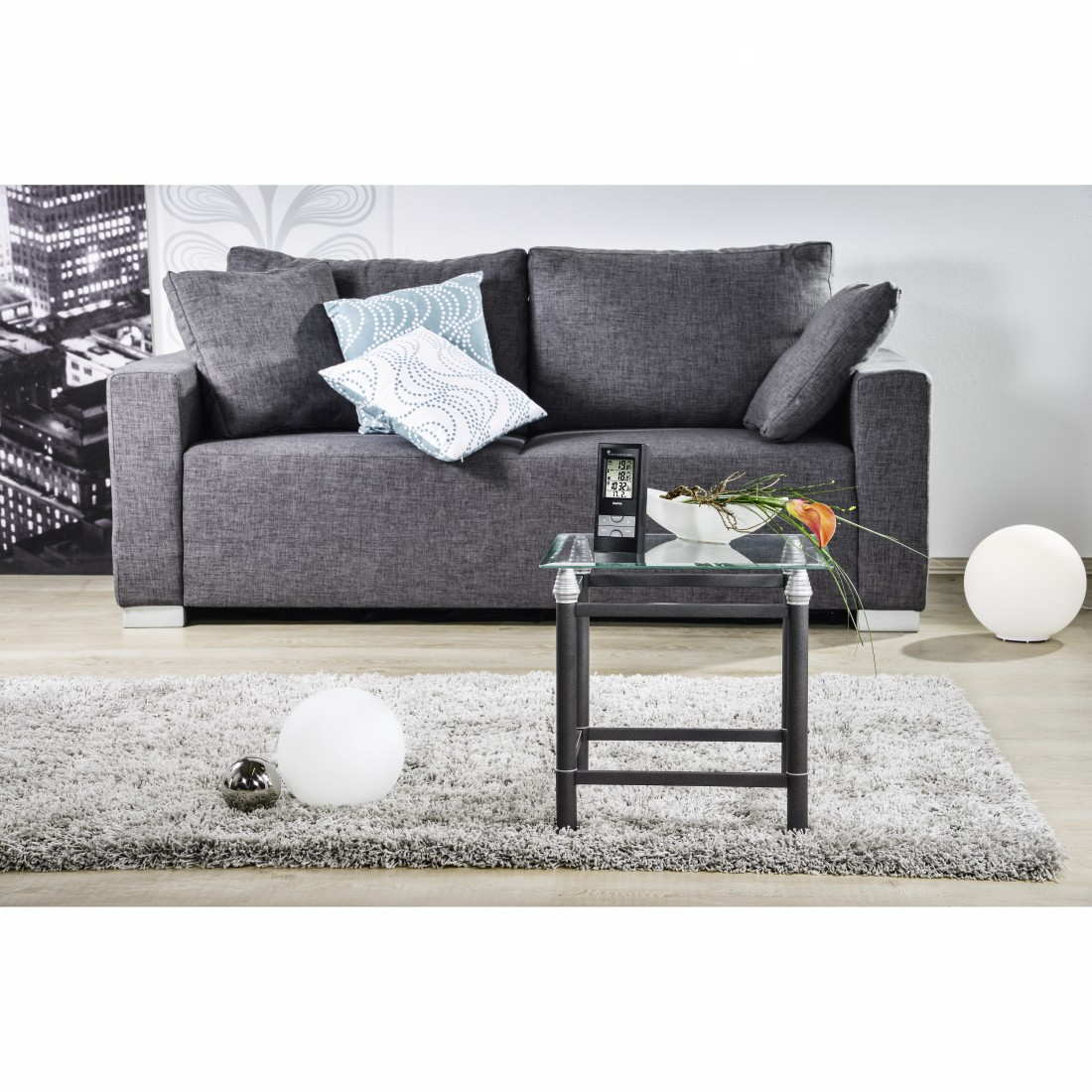 Sofá gris con almohadas, mesa de cristal con objetos en alfombra, dos lámparas de globo blancas.