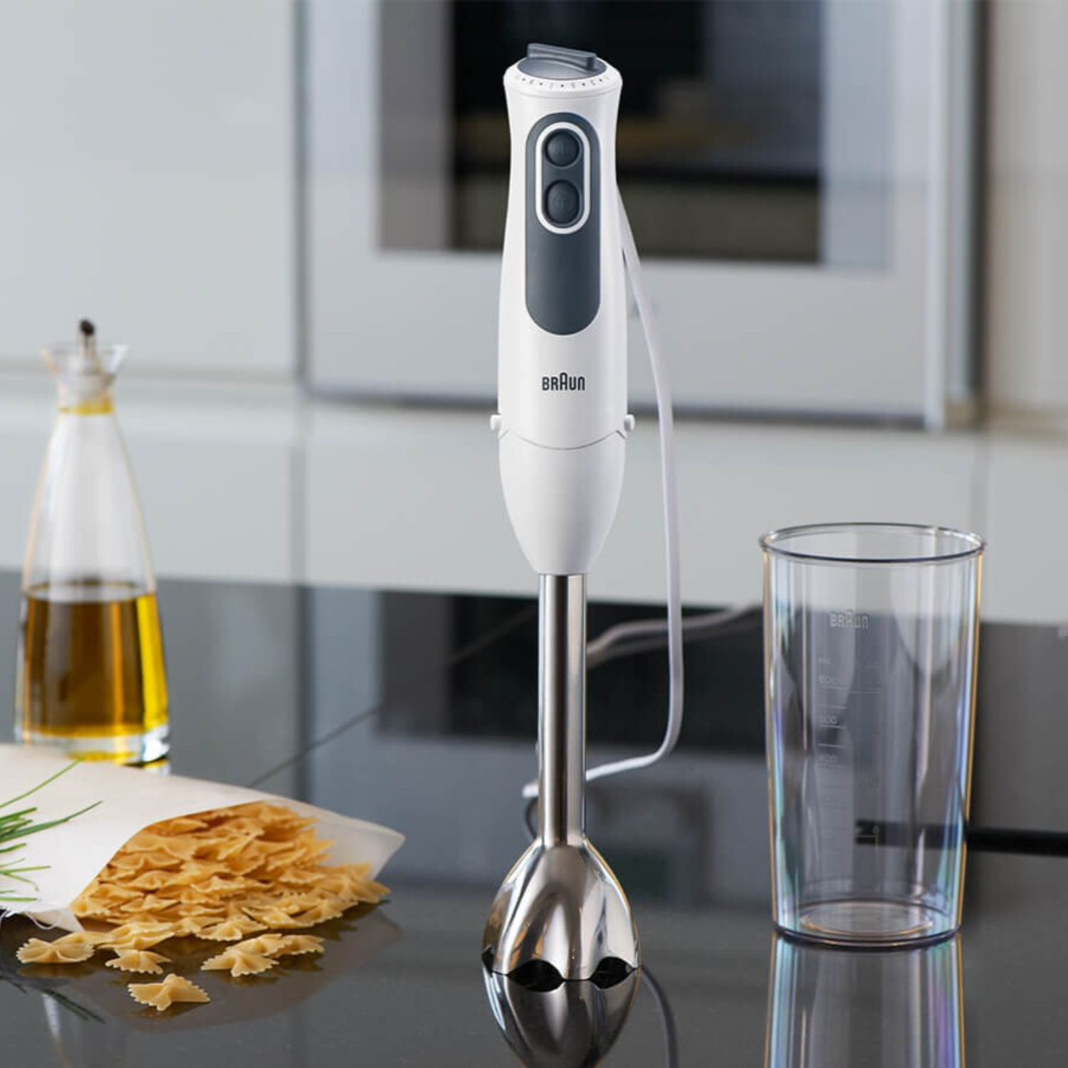 Witte en grijze staafmixer op zwart oppervlak met pasta en olie.