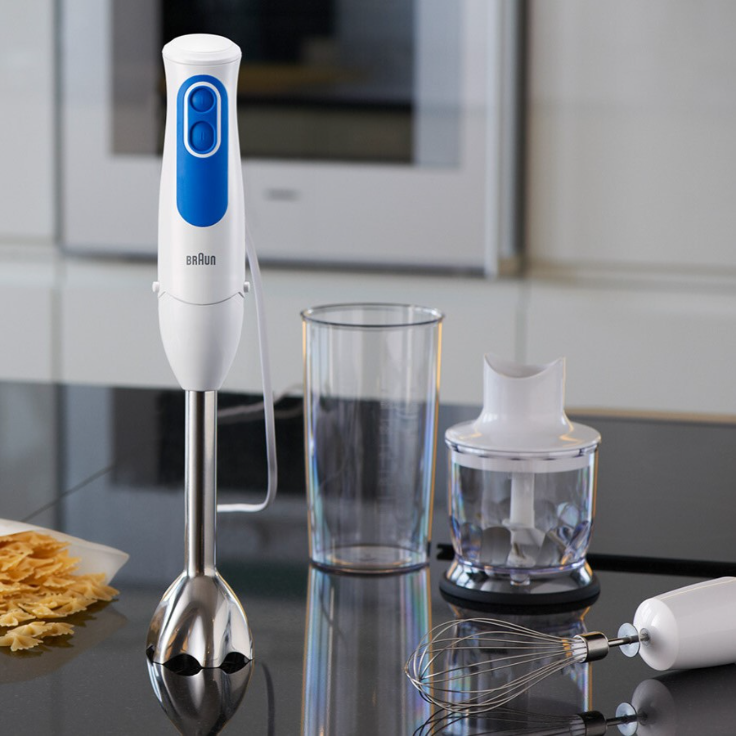 A white and blue Braun hand blender with attachments and a glass. The blender is on a black surface.