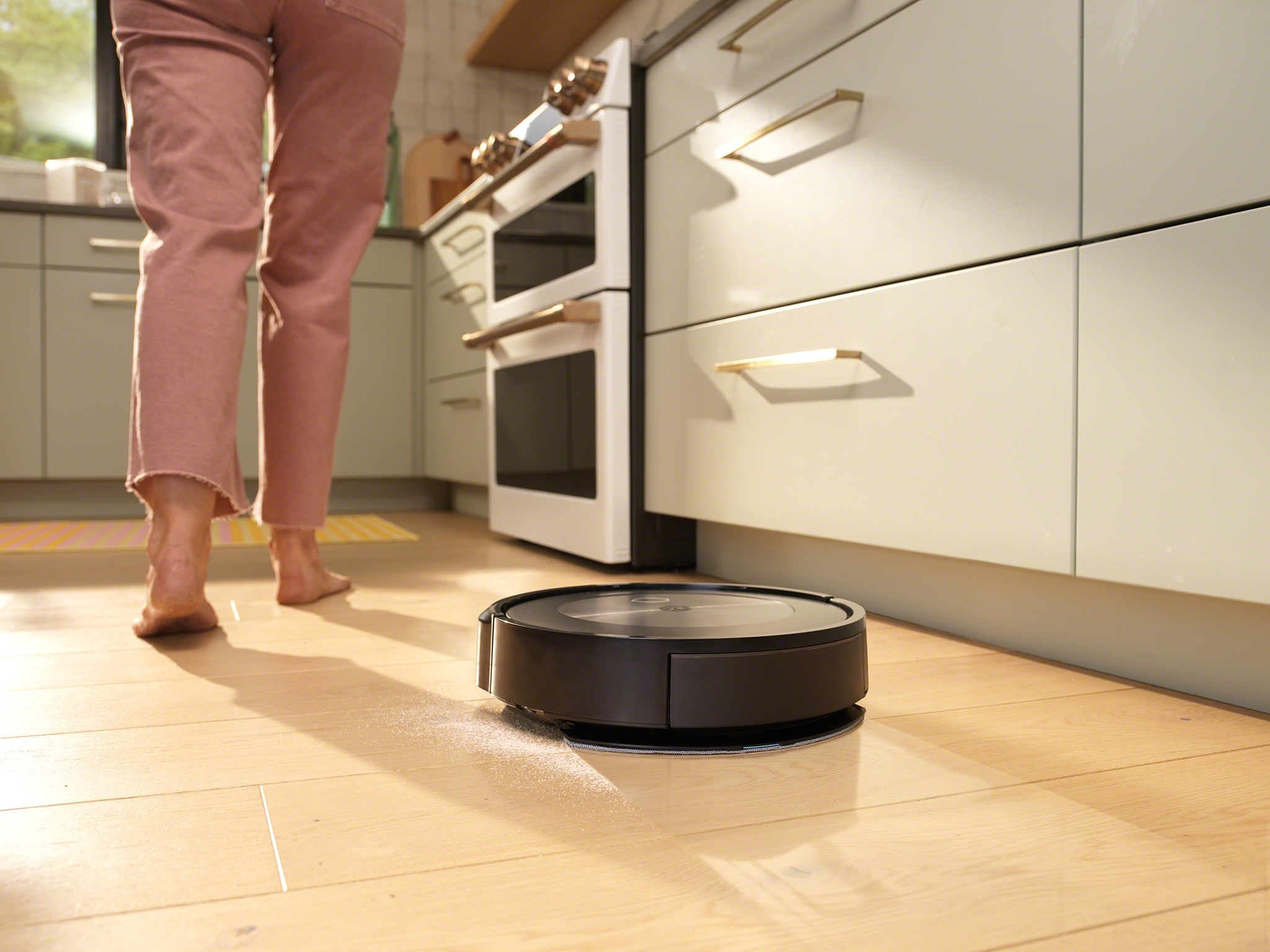 Ein Roboter-Staubsauger reinigt einen Holzboden. Eine Person geht im Hintergrund.