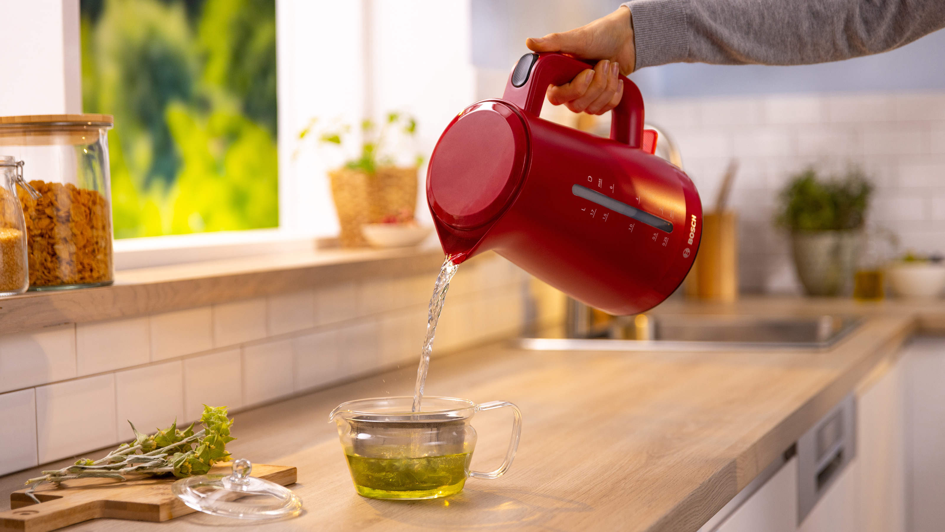 Ein roter Wasserkocher gießt Wasser in eine gläserne Teekanne mit Tee auf einer Holztheke.