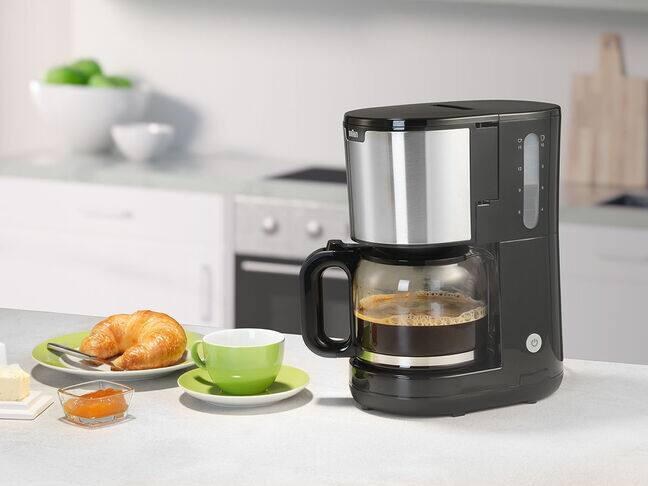 Eine schwarze Kaffeemaschine mit Kaffee im Glaskrug. Croissant, Butter und Marmelade auf einem Teller daneben.