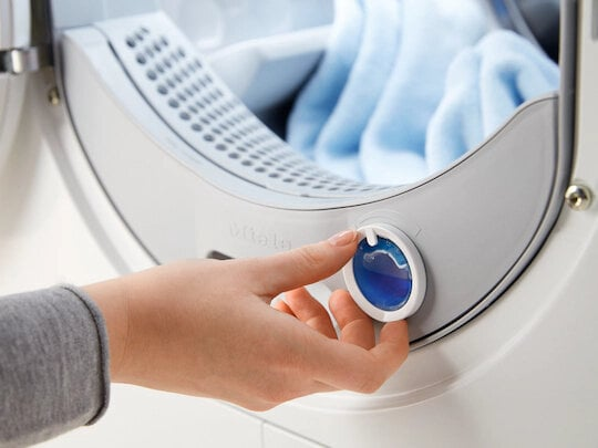 Ręka obraca niebieskie pokrętło na białej maszynie. W środku niebieskie ubrania.