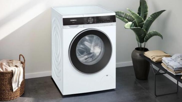 Biała pralka obok rośliny i kosza. Suszarka jest biała.