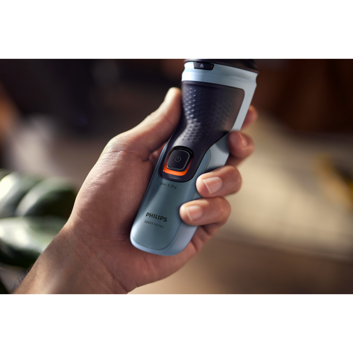 Hand holding a Philips electric shaver. It's blue and black with an orange power button.