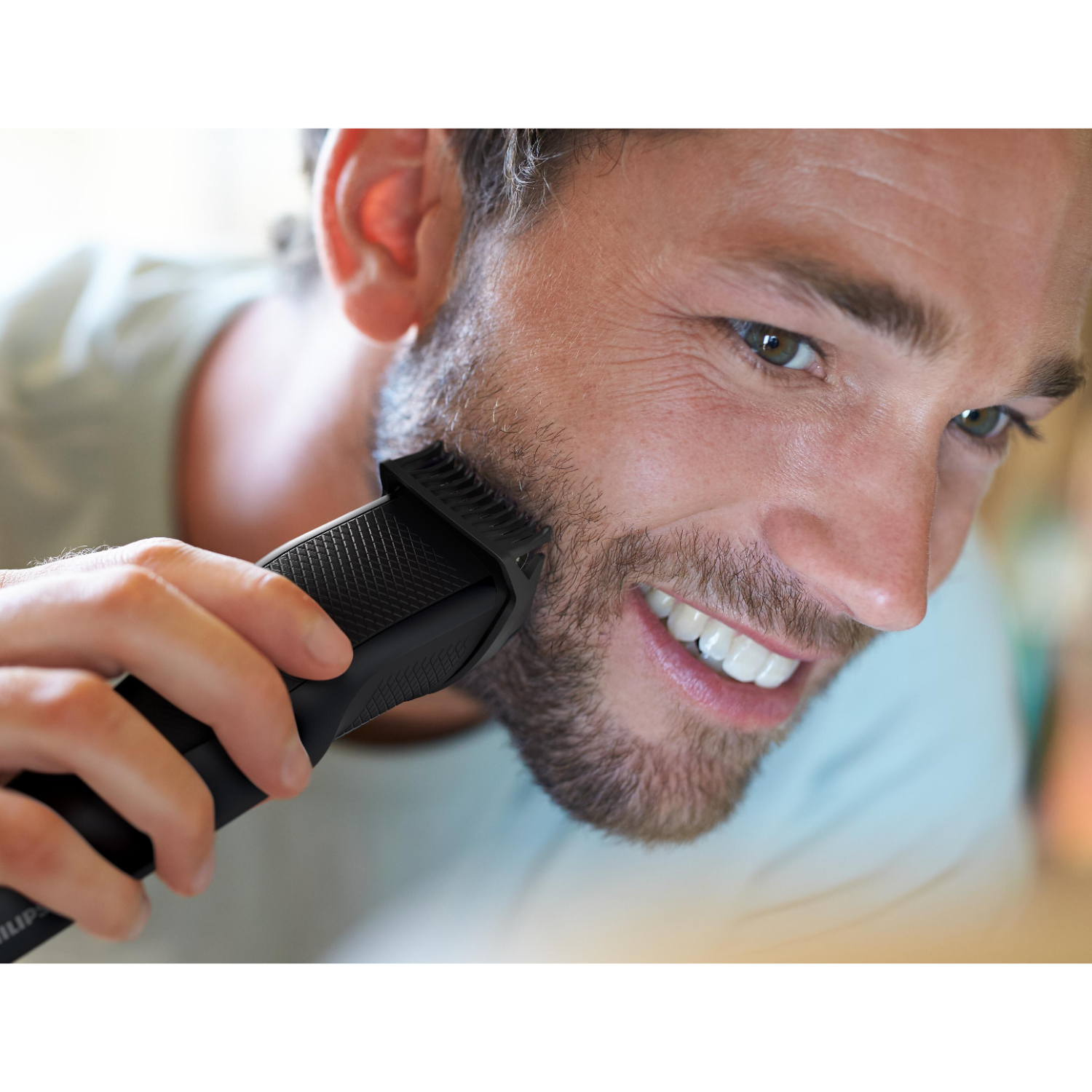 Man shaving his beard with a black trimmer. He's smiling, light skin, with a light blue shirt.