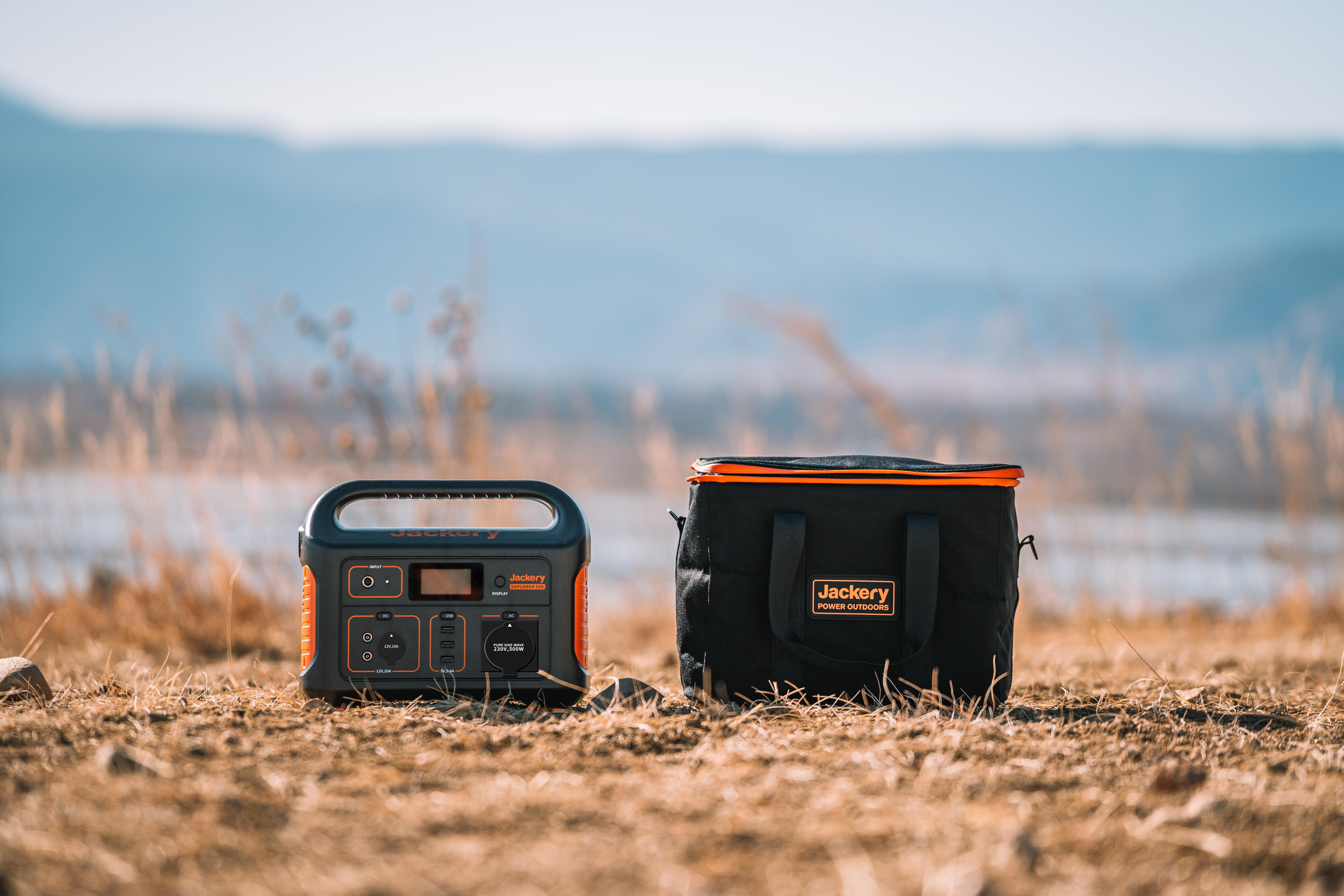 Zwei Jackery-Powerstations und eine schwarze Aufbewahrungstasche liegen am Boden. Ein Feld und Berge im Hintergrund.