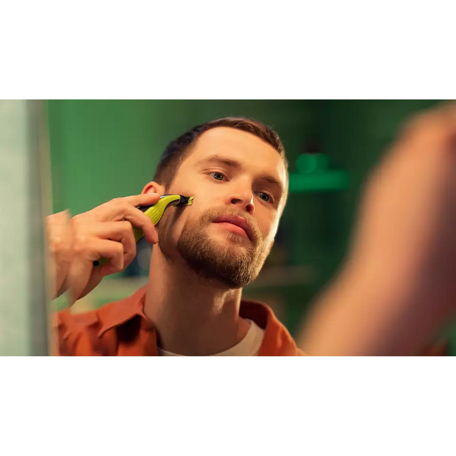 Man shaves his beard in the mirror. He holds a yellow and black trimmer. Green wall behind him.