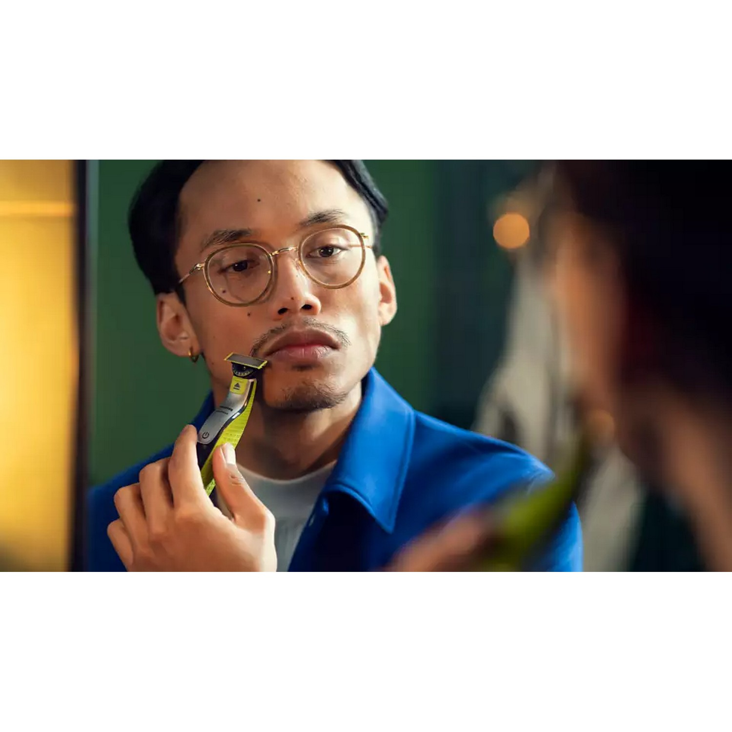 Man shaving his mustache in a mirror, using a green and black trimmer.