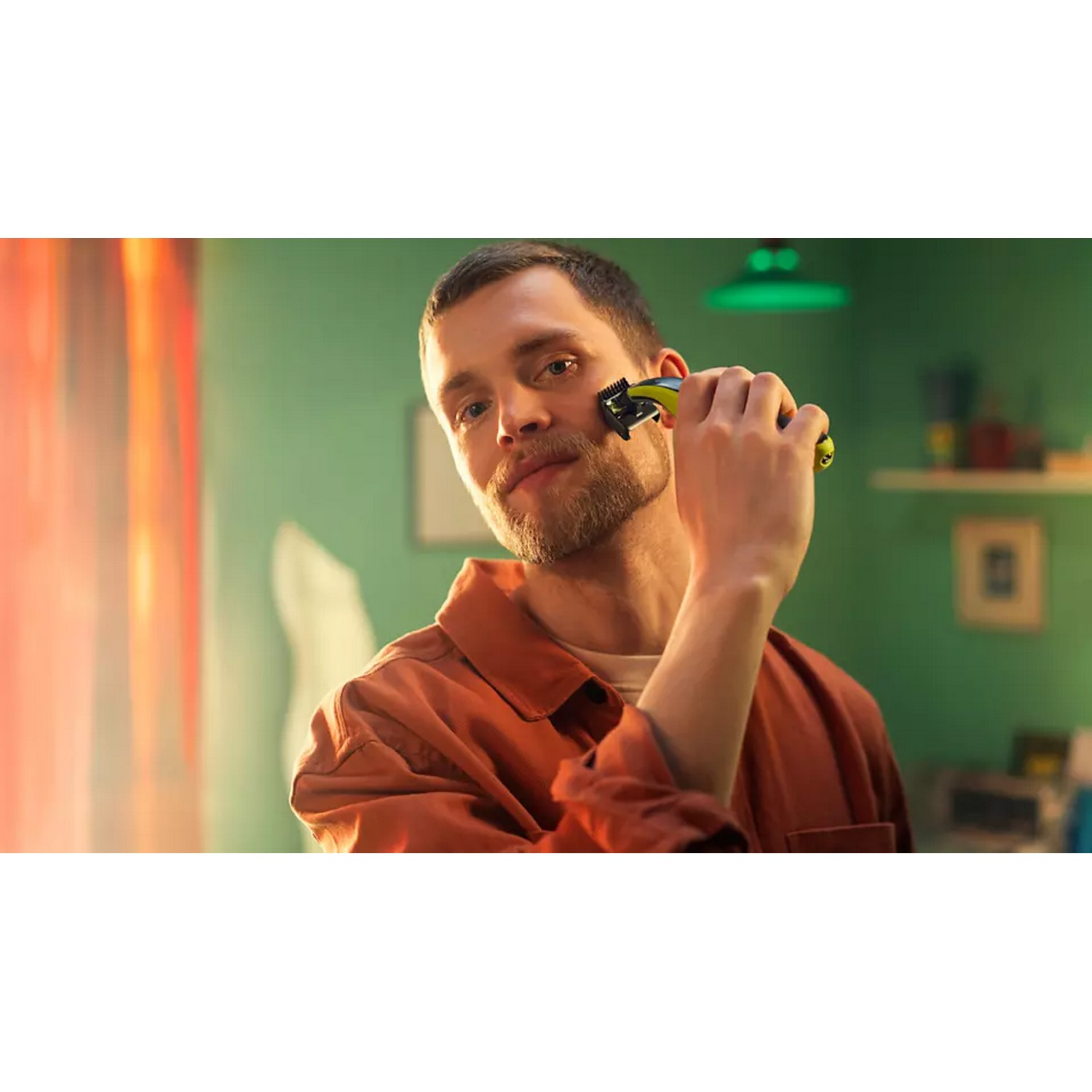 Man shaving his face with a blue and black trimmer, with a green wall background.