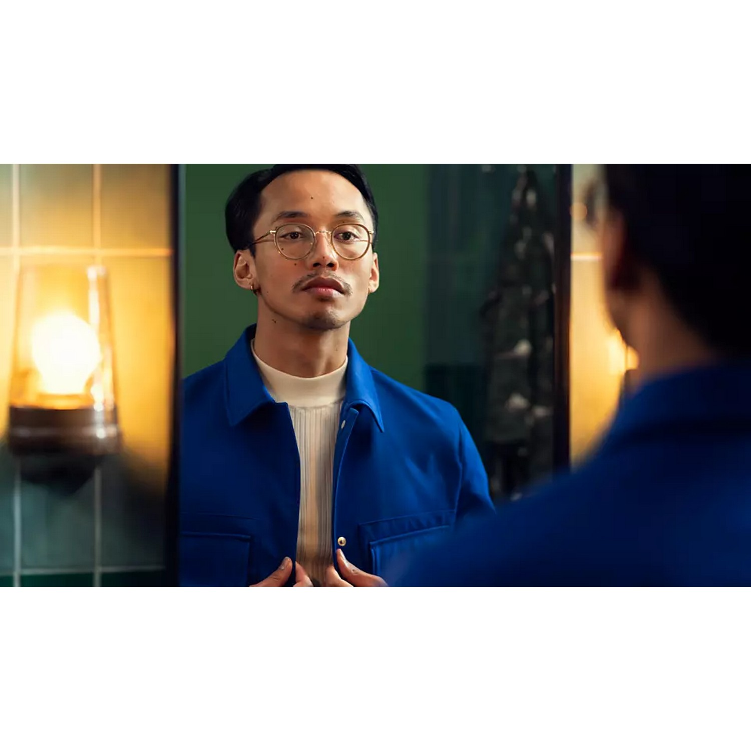 A man in a blue jacket looks at himself in a mirror, with a background of a bathroom.