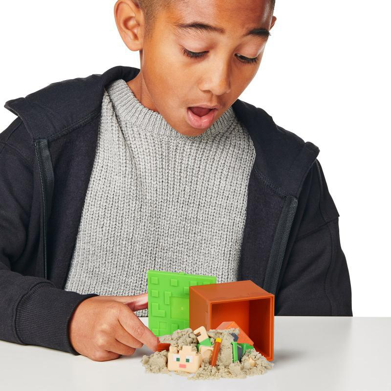 Junge betrachtet Minecraft-Spielzeug mit Sand, einem grünen Block und einer orangefarbenen Box.