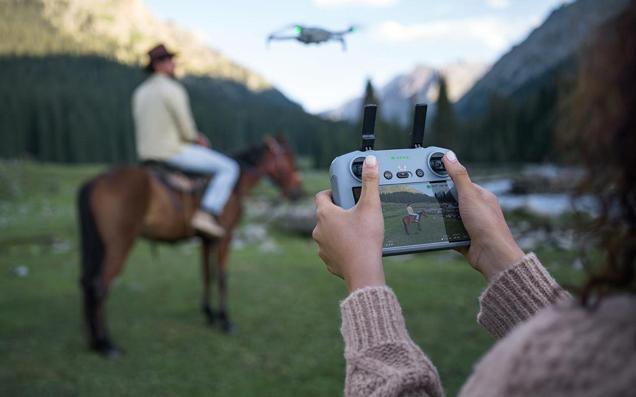 Person fliegt Drohne über ein Feld mit einer Person auf einem Pferd. Hügel und Bäume im Hintergrund.