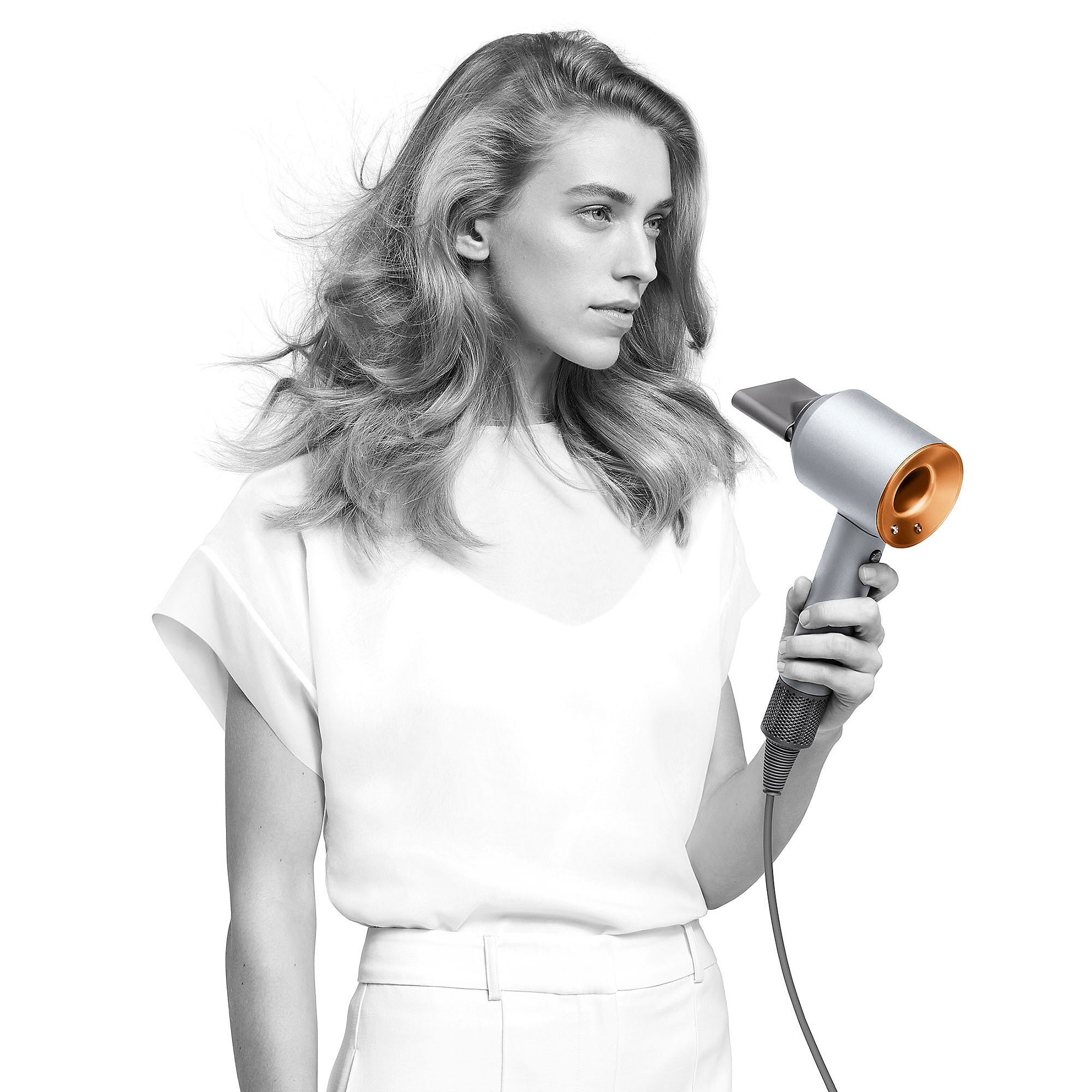Kobieta używa srebrnej suszarki do włosów ze miedzianymi akcentami. Włosy powiewają. Białe tło.