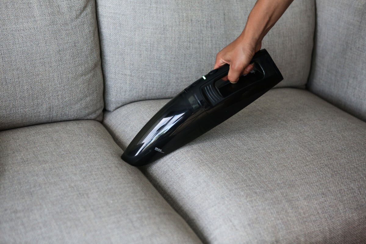 Ręka odkurza szarą sofę, używany jest czarny odkurzacz.
