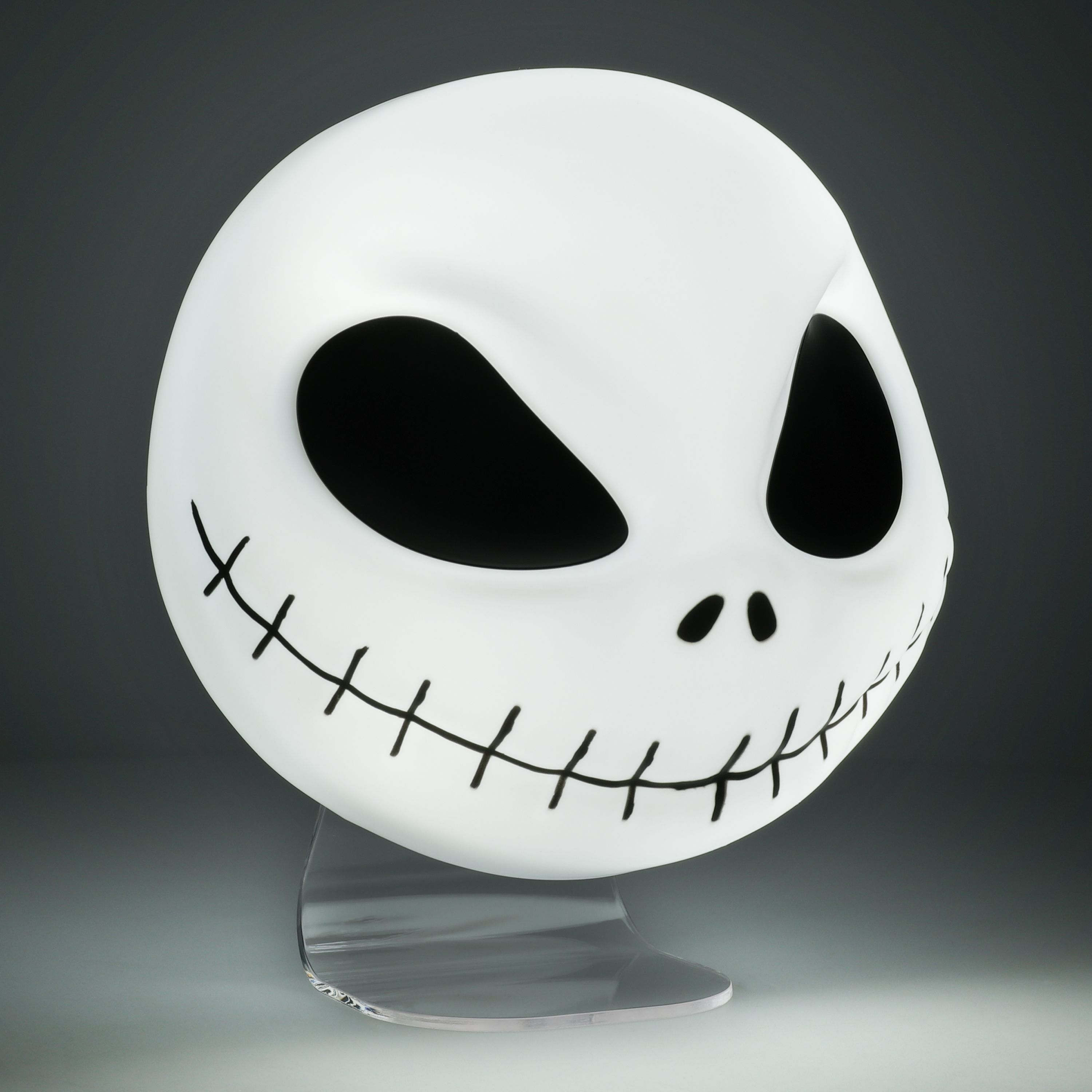 Jack Skellington Maske, weiß mit schwarz genähtem Mund, auf einem klaren Ständer, auf dunklem Hintergrund.