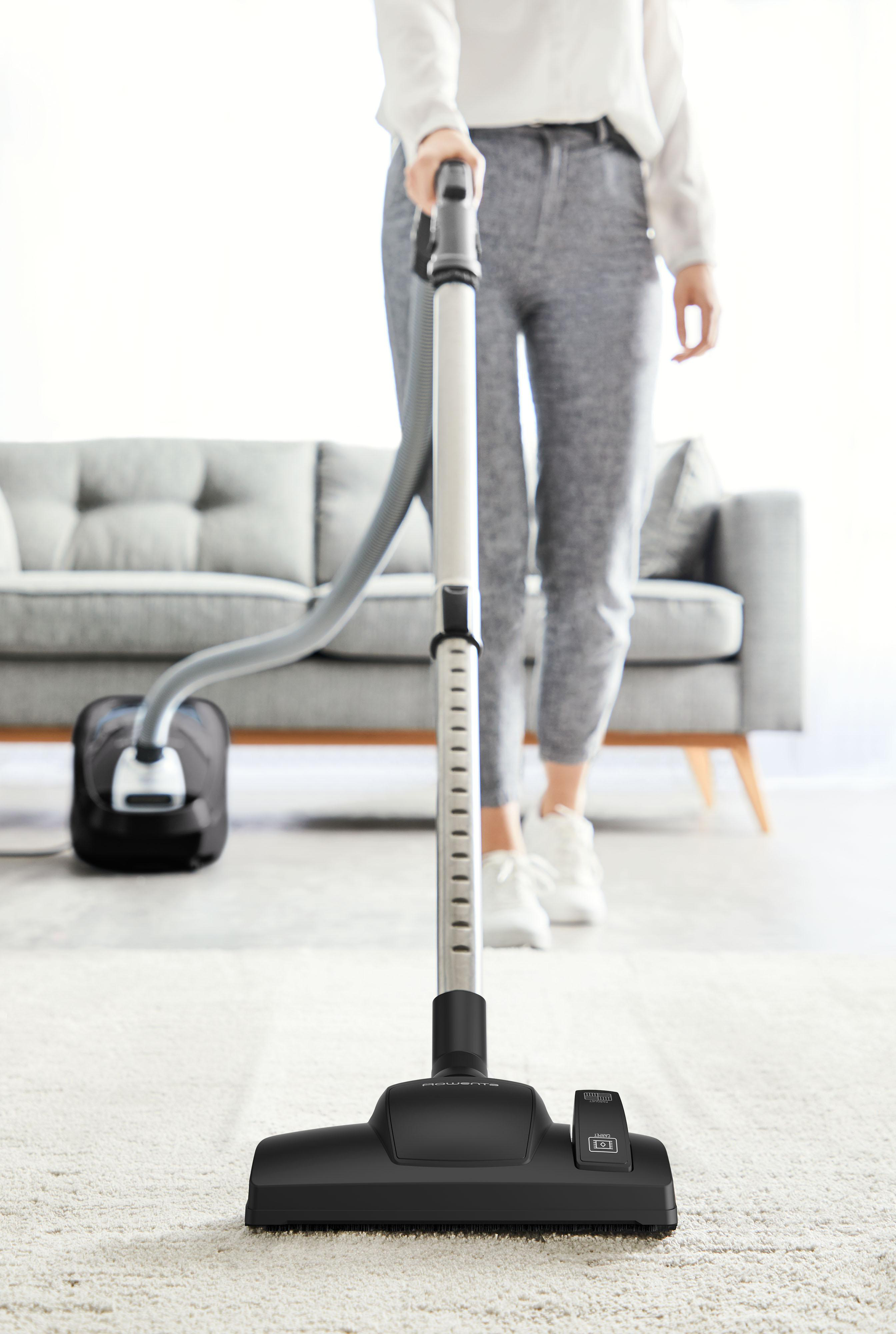 Person saugt Teppich mit schwarzem Staubsauger, graues Sofa im Hintergrund.