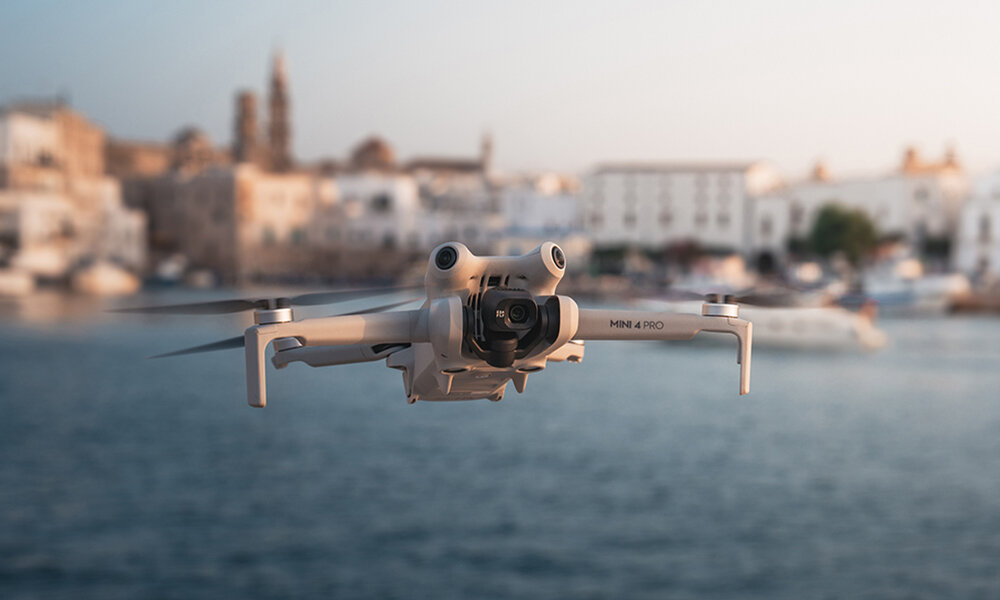 Szary dron lecący nad wodą z rozmytymi budynkami w tle. Dron jest skierowany w stronę widza.