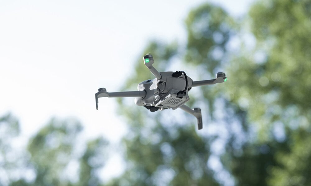 Dron latający w powietrzu z zielonymi drzewami w tle.