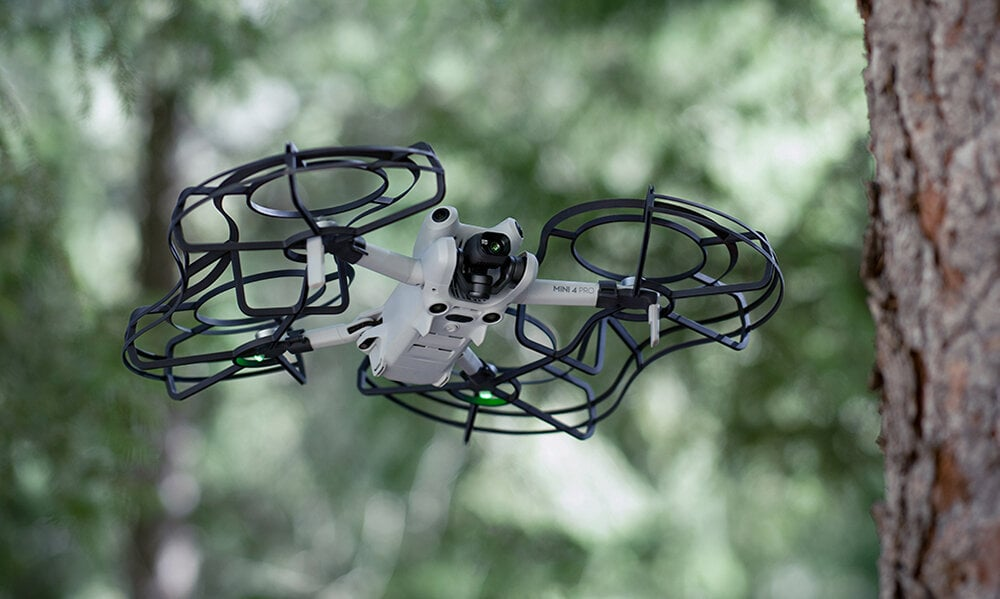 Biały dron z czarną klatką latający blisko pnia drzewa, rozmyte zielone tło.