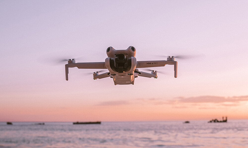 Dron w locie nad wodą o zachodzie słońca, różowe i pomarańczowe niebo, plaża w tle.