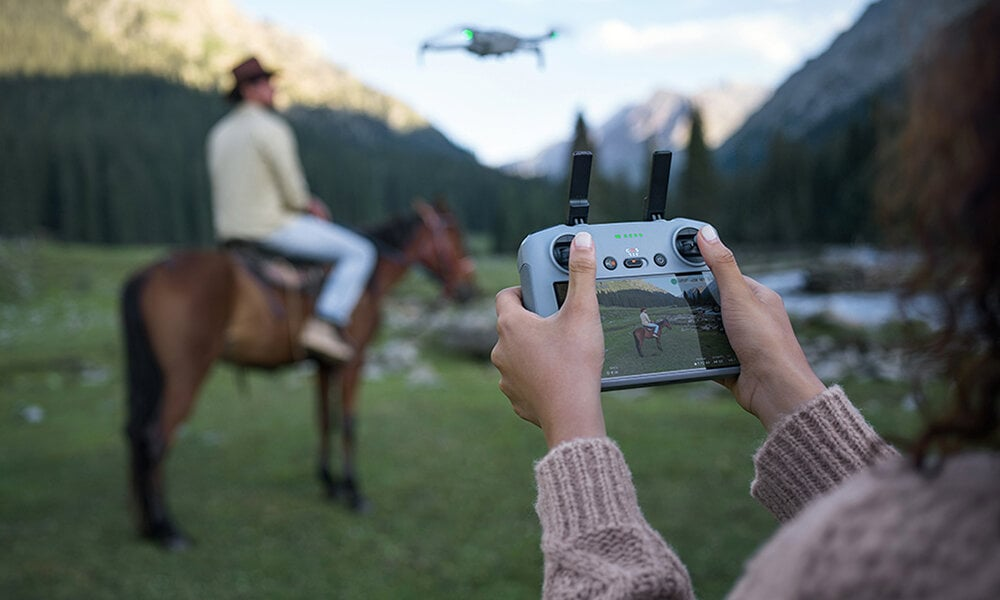 Osoba steruje dronem za pomocą pilota. Kawałer na koniu w tle.