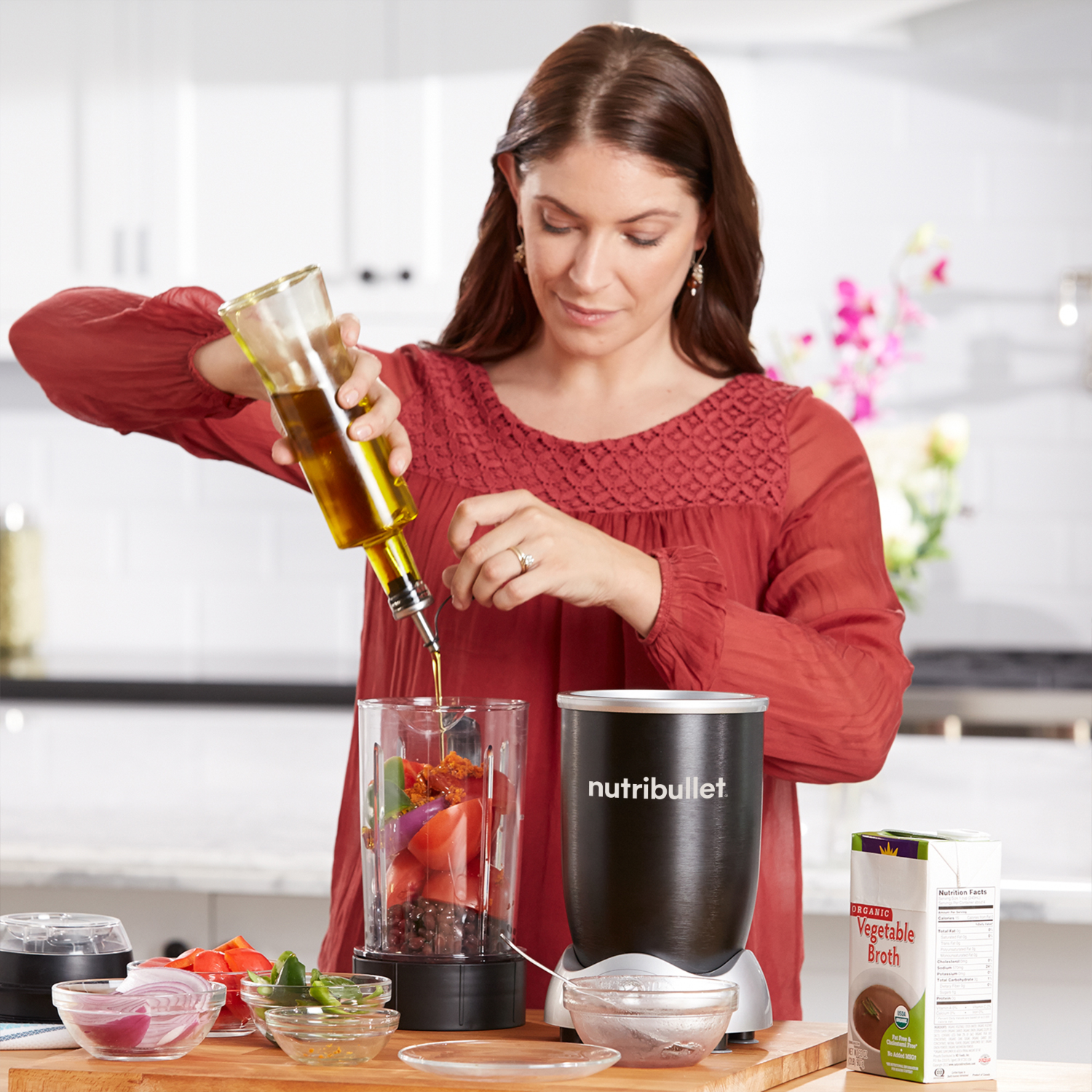 A woman pours oil into a blender with vegetables, preparing a meal in a bright kitchen.