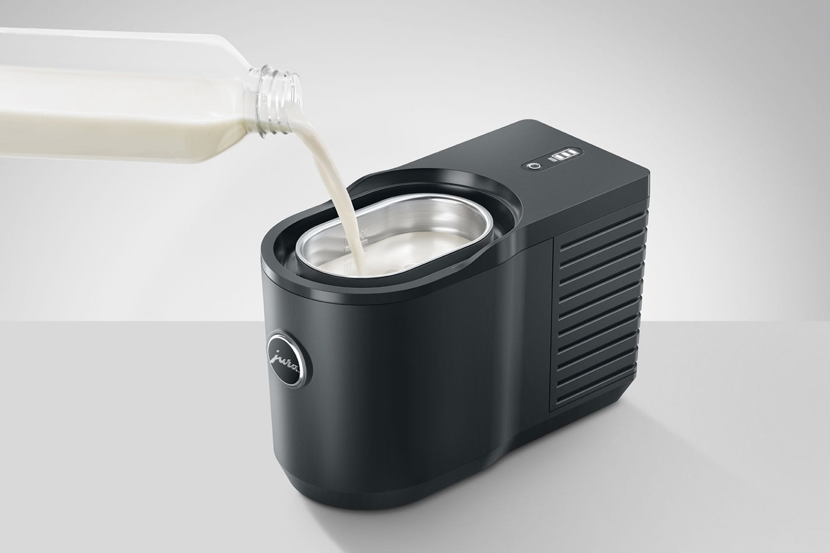 Milch wird in einen schwarzen Milchaufschäumer gegossen. Milchflasche links oben.