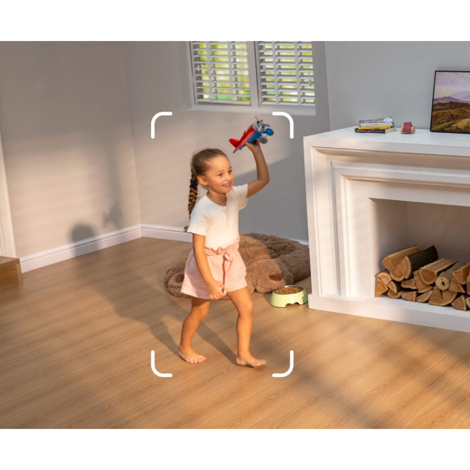 A girl holding a toy airplane. She is in a room with a fireplace and wooden floor.