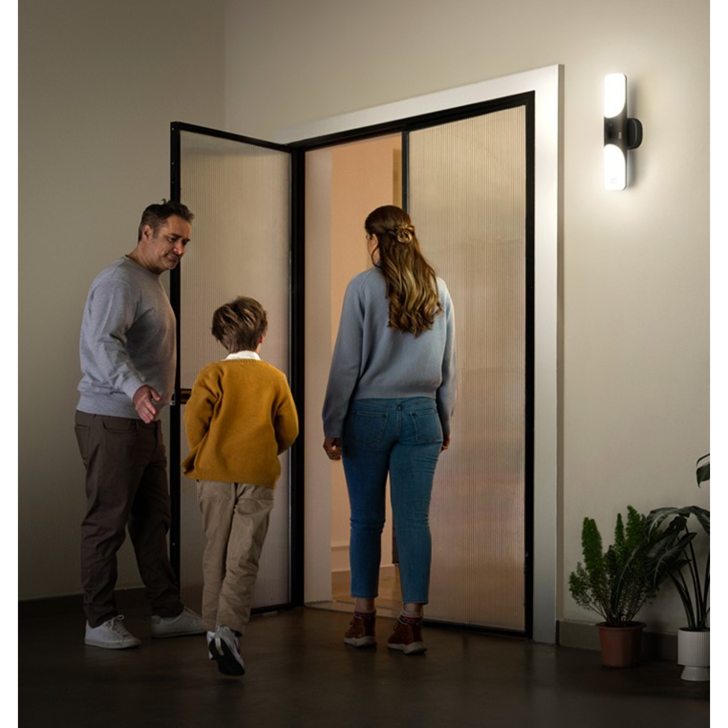 A family entering a home through a glass door, with a wall-mounted light fixture.
