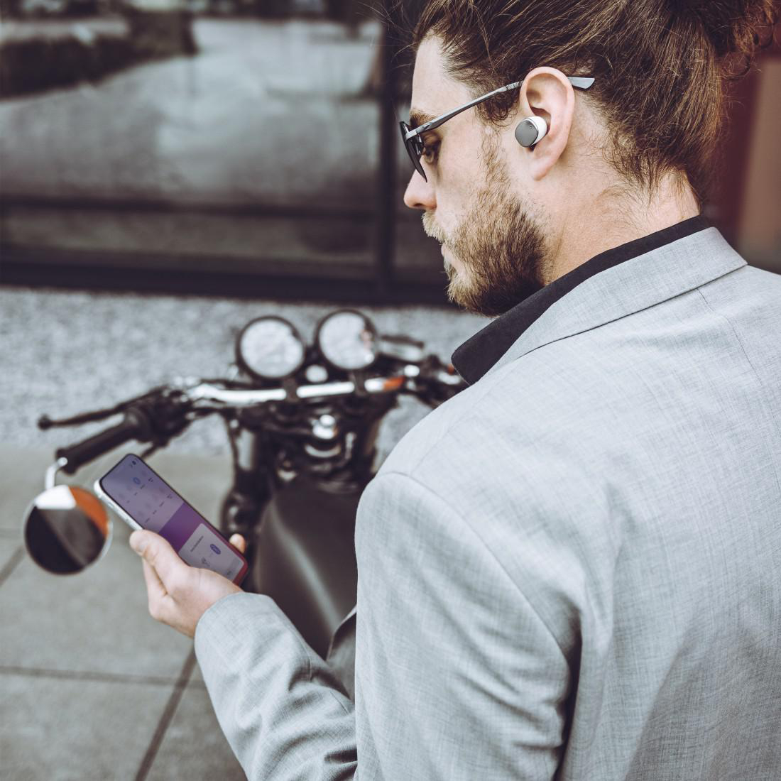 Mann mit Ohrhörern, Telefon in der Hand, in der Nähe eines Motorrads. Hintergrund: Stadt.