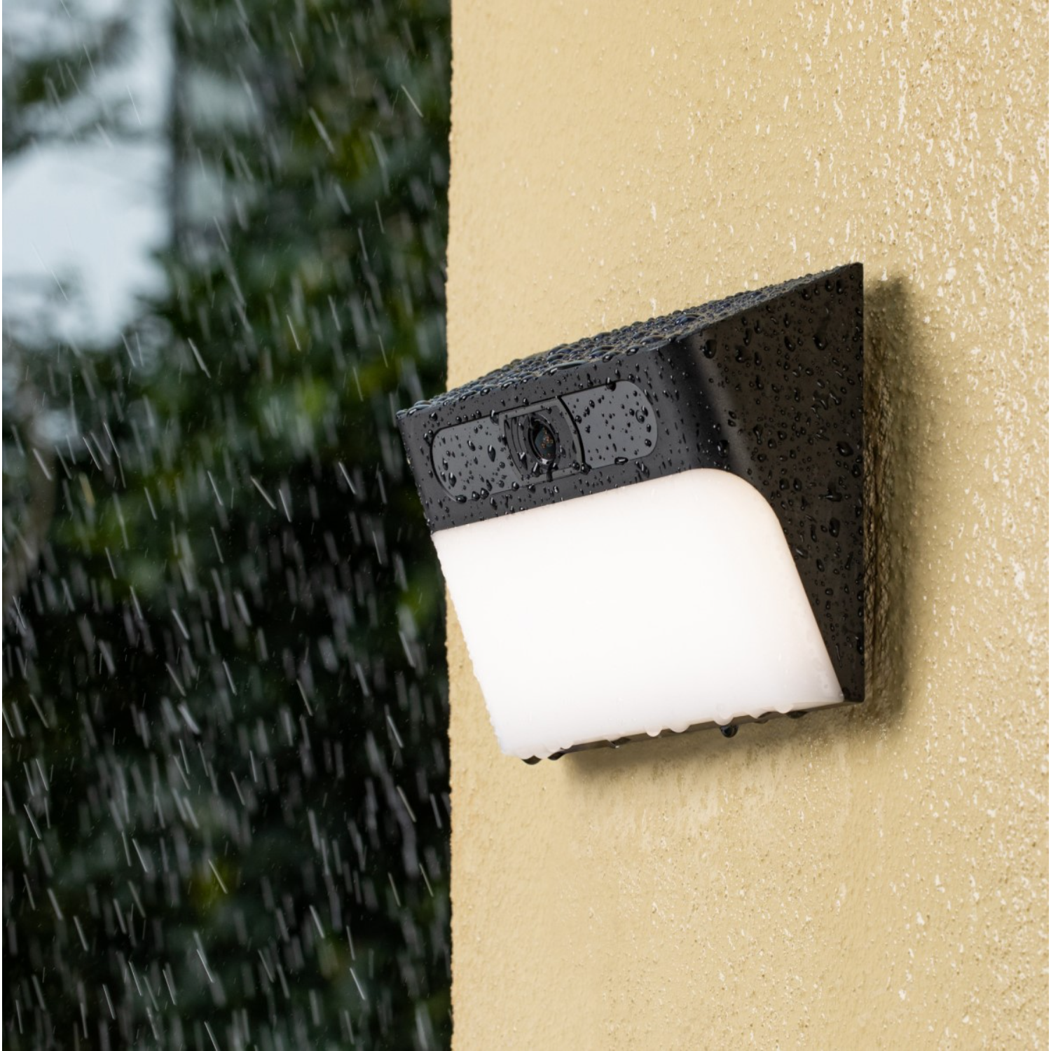 Zwarte buitenverlichting op een beige muur, regen valt. Het licht is wit.
