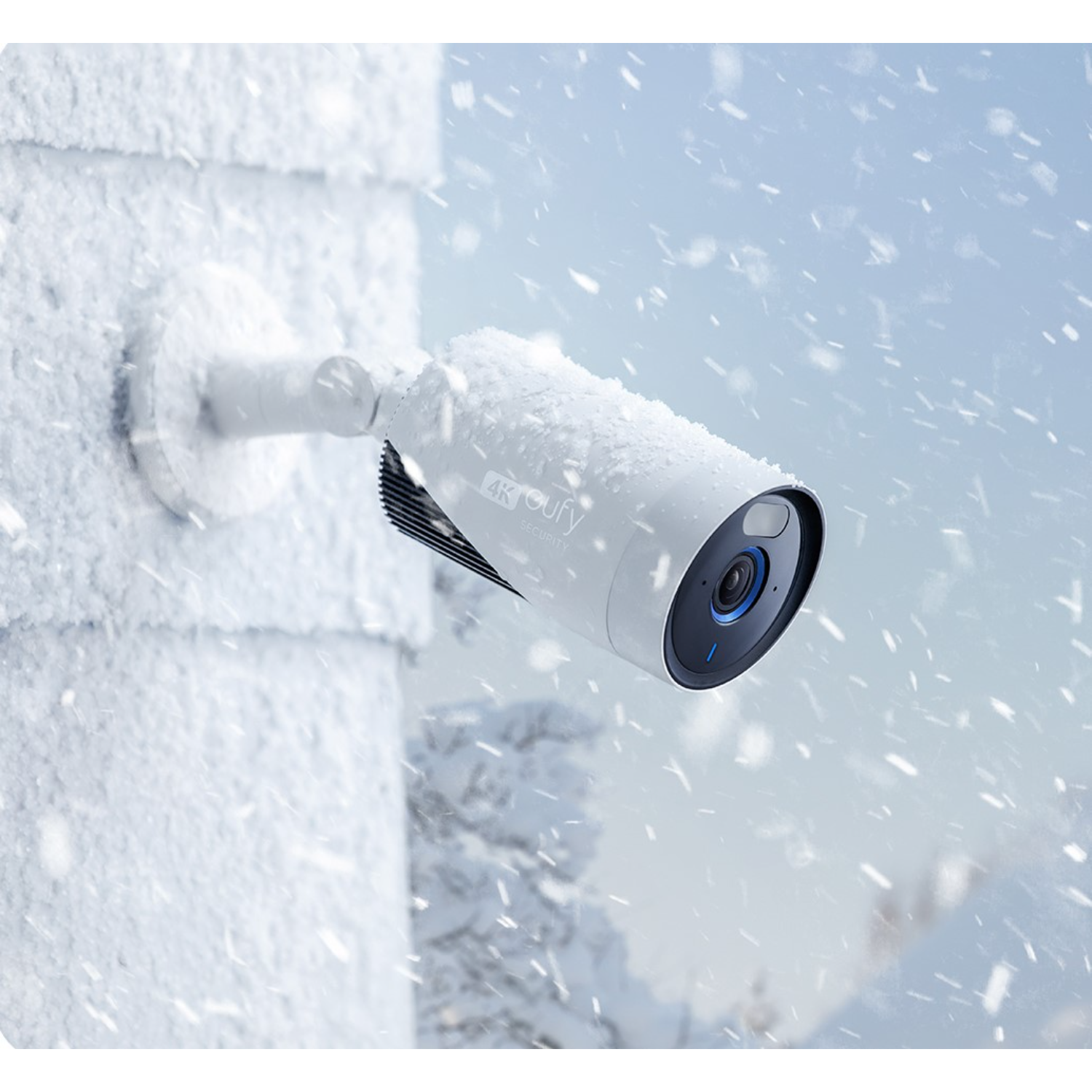 A white security camera covered in snow, mounted on a white wall. Snow is falling.
