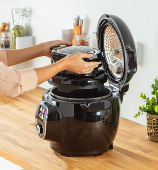 Ręce umieszczają czarną pokrywę na czarnym szybkowarze. Kuchnia w tle.