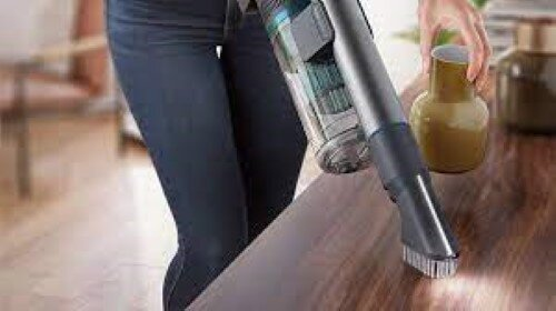 Osoba odkurzająca drewnianą powierzchnię. Widoczny odkurzacz i słoik.